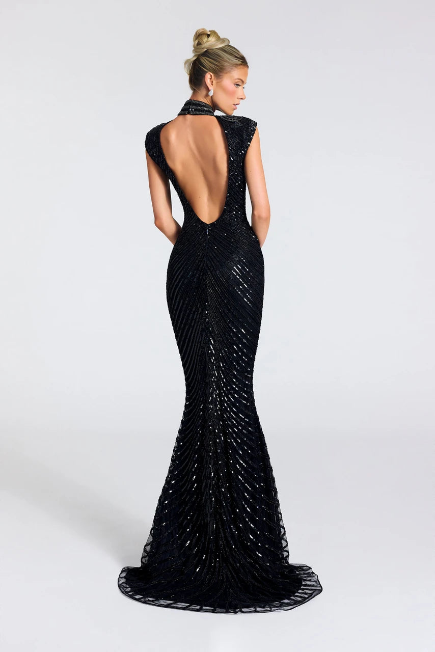 A woman modeling a black floor-length evening gown from the back, featuring a deep open back, cap sleeves, and dense beaded vertical detailing that creates a shimmering effect, set against a plain light gray studio background.
