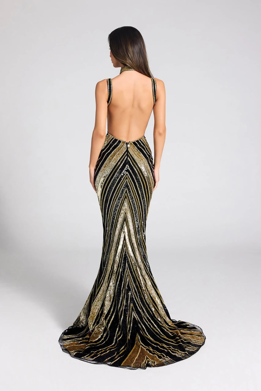 A woman viewed from the back wearing a floor-length open-back evening gown with a metallic chevron pattern in black, gold, and bronze tones, photographed against a light gray studio background.