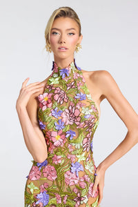 A model wearing a sheer halter-neck dress with vibrant 3D floral applique in pink, purple, green, and yellow on a light gray studio background.
