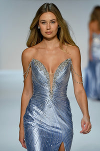 A model walks a fashion runway wearing a strapless, shimmering silver gown with intricate beadwork, a plunging V-neckline, and delicate chain details on the shoulders.