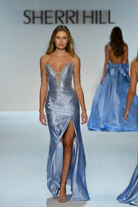 A model walks the runway wearing a shimmering light blue evening gown with a plunging neckline, beaded embellishments, and a thigh-high slit. The Sherri Hill brand name is visible on the runway backdrop, with another model in a matching blue gown in the background.