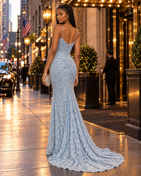 A woman stands on a city sidewalk at dusk wearing a light blue lace mermaid gown with a fitted bodice, ruched back, and long train. The background shows a lit hotel entrance, street lamps, and pedestrians.