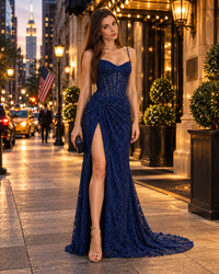 A woman stands on a city sidewalk at dusk wearing a deep navy lace evening gown with thin straps, a fitted bodice, a high thigh slit, and a long train. The background shows illuminated buildings, streetlights, and city traffic.