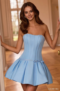 A woman modeling a strapless light blue mini dress with a corset-style bodice, pearl-like embellishments along the neckline, and a flared skirt, posed indoors with her arms outstretched.