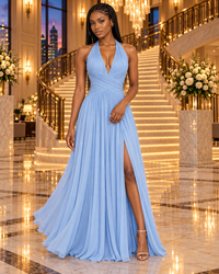 A woman models a light blue halter-neck floor-length gown with a deep V-neckline, pleated bodice, and a high thigh slit, set against a luxurious hotel lobby with a grand illuminated staircase.
