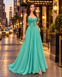 A woman stands on a city sidewalk at dusk wearing a floor-length mint green gown with a sweetheart neckline, bow at the bust, and a full flowing skirt.