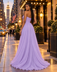 A woman stands on a city sidewalk wearing a strapless lilac color formal gown with a fitted bodice and full flowing skirt. The background shows a lit urban street scene at dusk with buildings, lamps, and pedestrians.