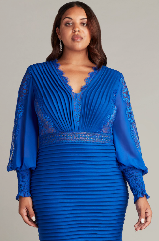 A woman wearing a knee-length pleated textured crepe cocktail dress with Bishop sleeves and a peekaboo lace hem, perfect for elegant evening occasions.