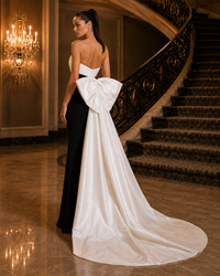 A woman stands on a grand staircase landing wearing a formal gown with a black body and a large white bow at the back, photographed indoors with a chandelier and staircase in the background.