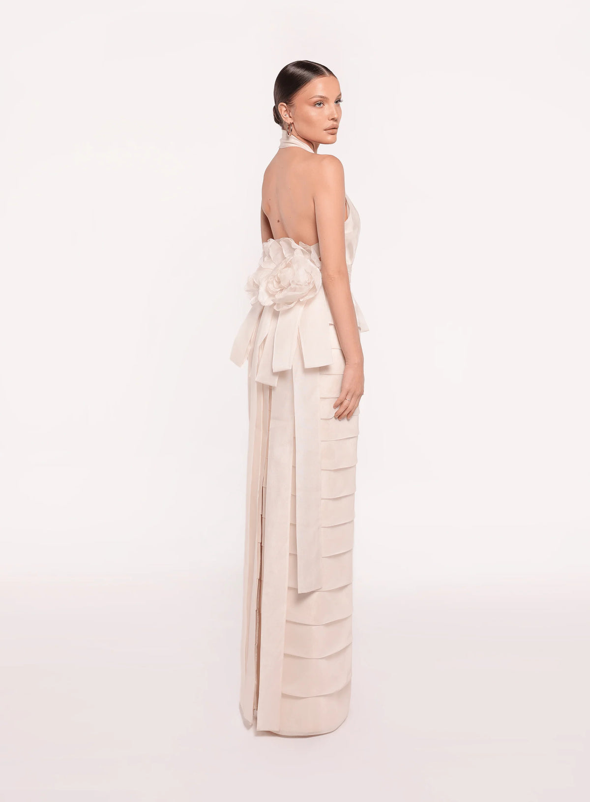 Model wearing the Tarik Ediz 98819 halter plunge neckline column gown with tiered skirt detailing, open back, floral accent, and long sash.