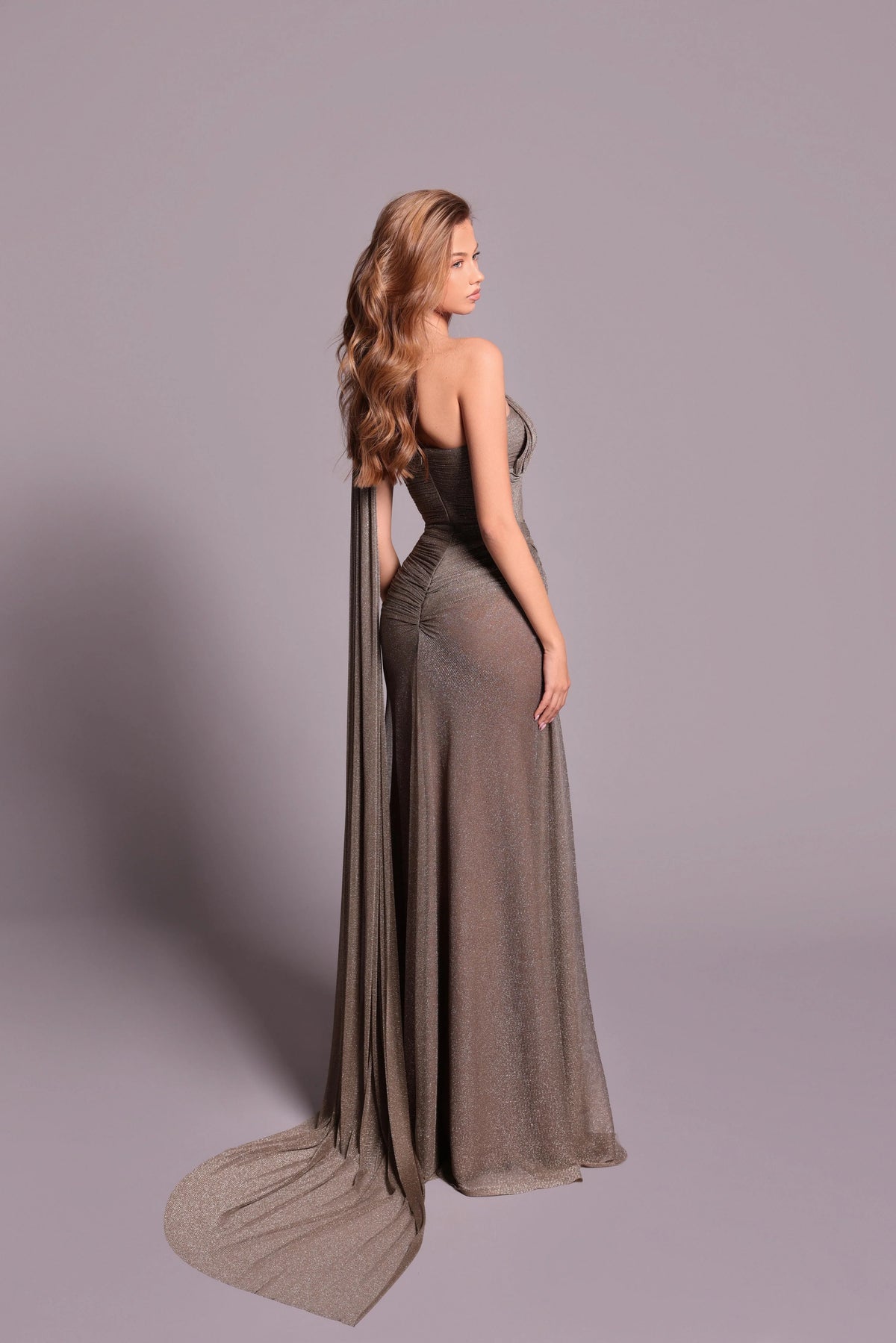 Model wearing Tarik Ediz 99041 one-shoulder draped gown with high slit and flowing back panel.