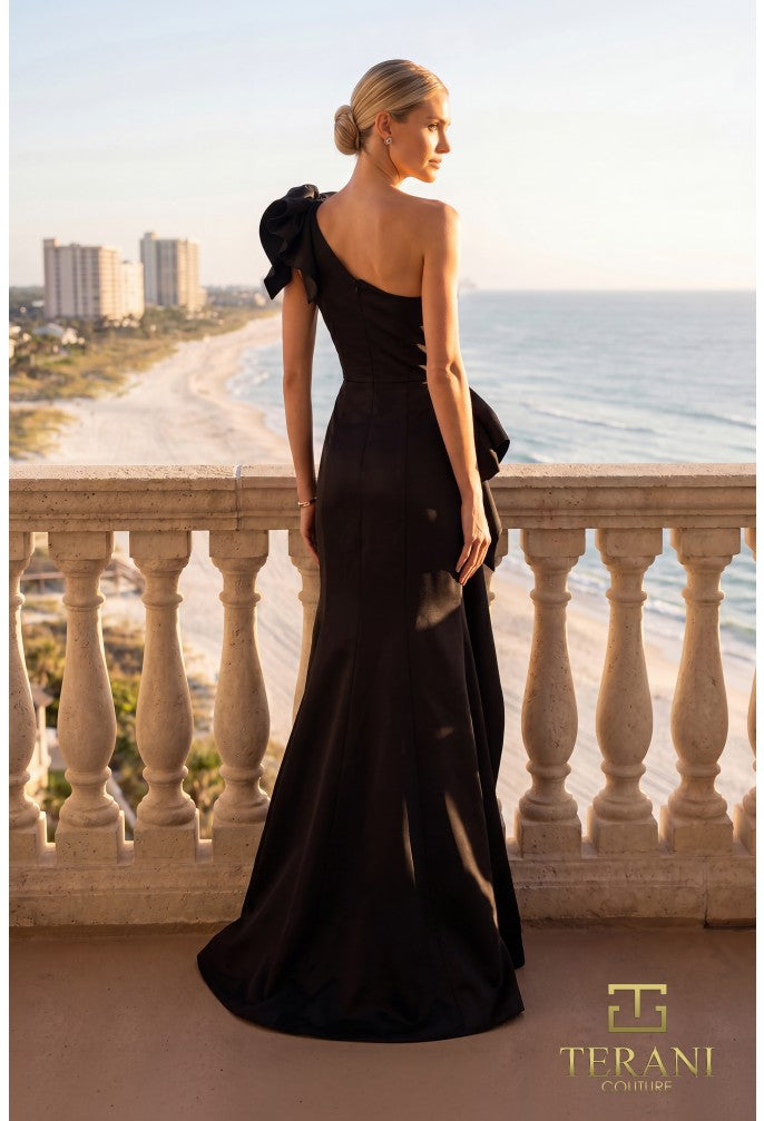Woman in a black one-shoulder dress standing on a balcony with a cityscape and ocean view, Terani Couture logo visible.