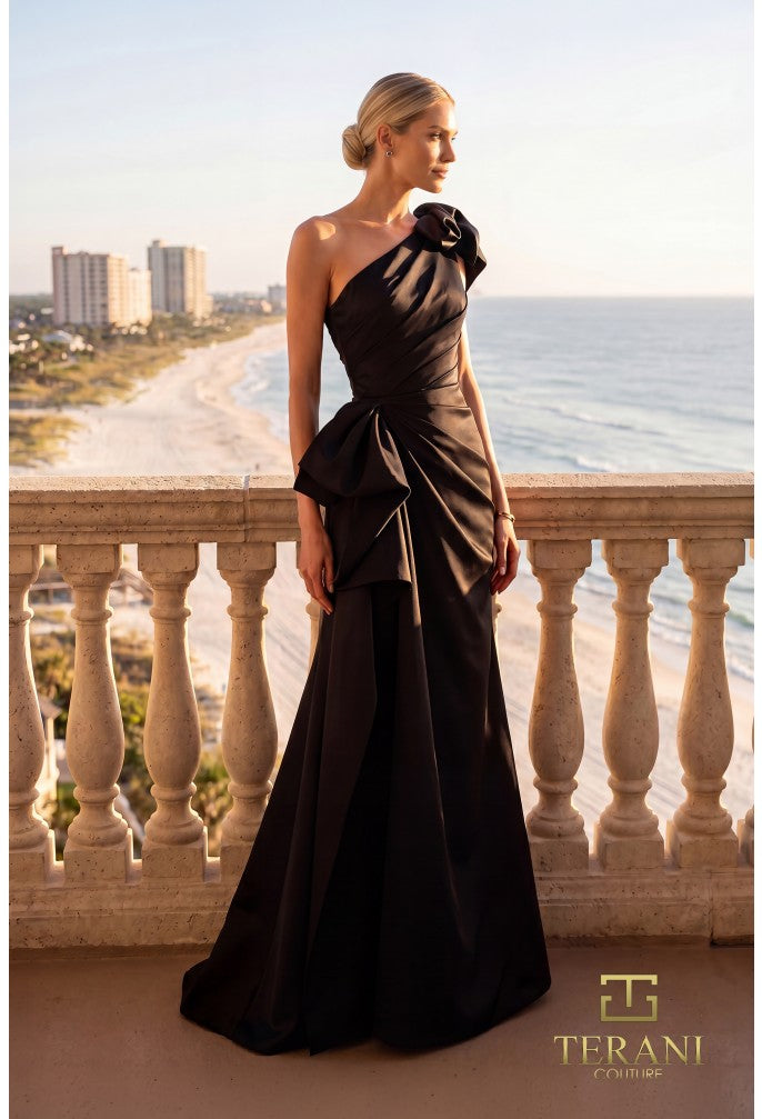 Woman in a black one-shoulder dress standing on a balcony with ocean view, Terani Couture logo visible.