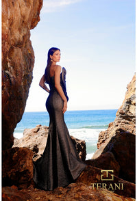 Woman in a elegant gown standing among rocks with ocean view, Terani brand visible