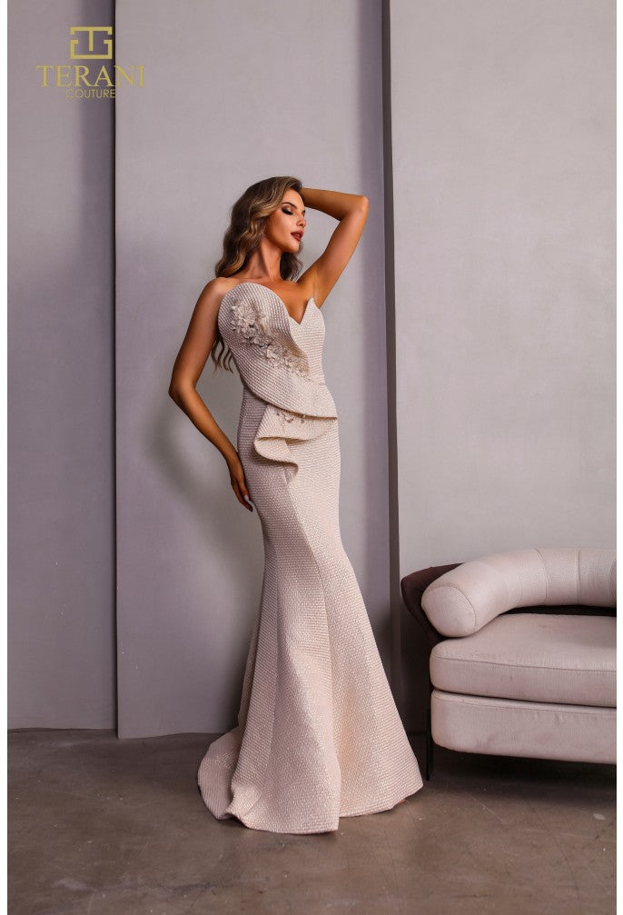 Woman in a white evening gown with lace details standing against a gray wall.