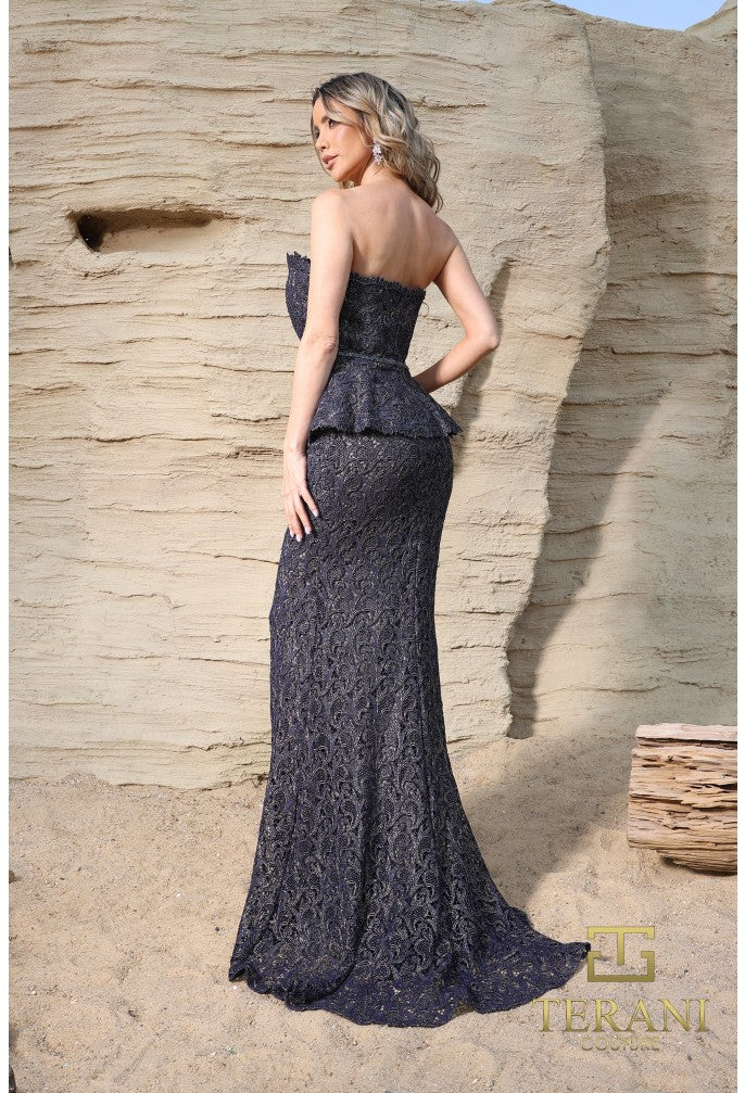 Woman in a strapless, floor-length dark blue lace dress standing against a textured beige wall.