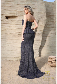 Woman in a strapless, floor-length dark blue lace dress standing against a textured beige wall.