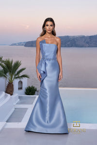 Woman in a light blue evening gown standing in front of a scenic backdrop with palm trees and a pool.