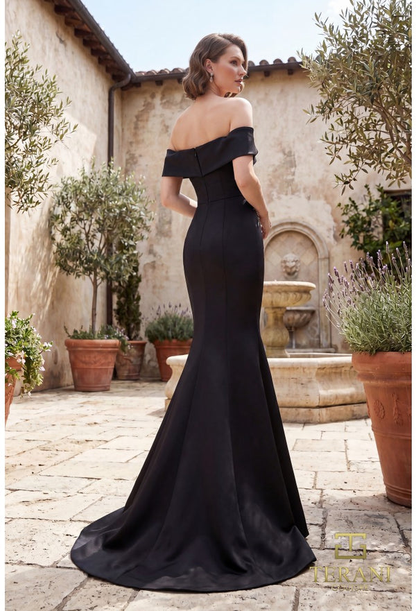 A woman wearing a black off-the-shoulder mermaid evening gown stands in a sunlit courtyard with stone walls, potted plants, and a fountain in the background.
