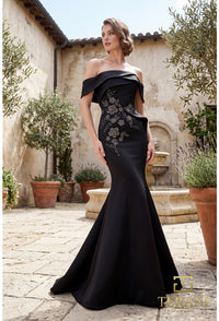 A woman models an off-the-shoulder black evening gown with floral embroidery on the bodice, set in a sunlit stone courtyard with potted plants and a rustic building in the background.