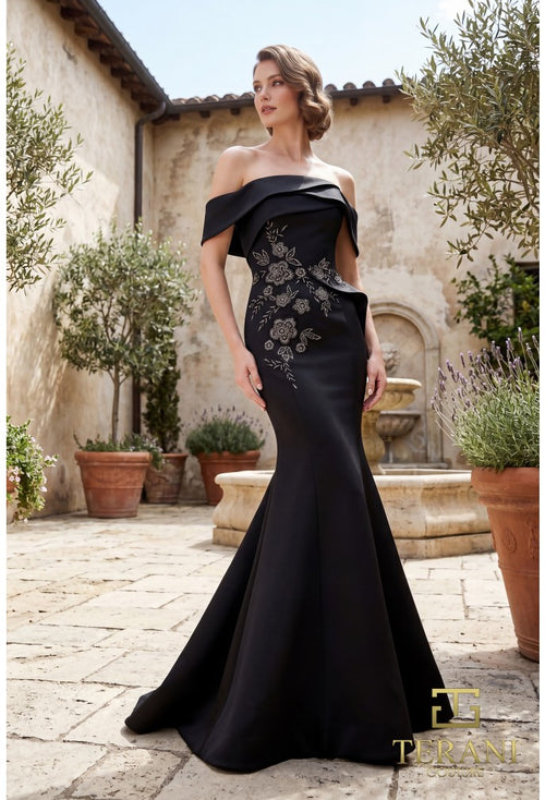 A woman models an off-the-shoulder black evening gown with floral embroidery on the bodice, set in a sunlit stone courtyard with potted plants and a rustic building in the background.