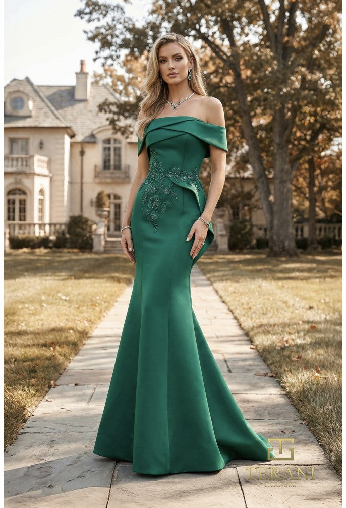 A woman stands on a stone path in front of a large house, wearing an off-the-shoulder emerald green mermaid gown with floral embroidery on the bodice.