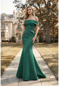 A woman stands on a stone path in front of a large house, wearing an off-the-shoulder emerald green mermaid gown with floral embroidery on the bodice.