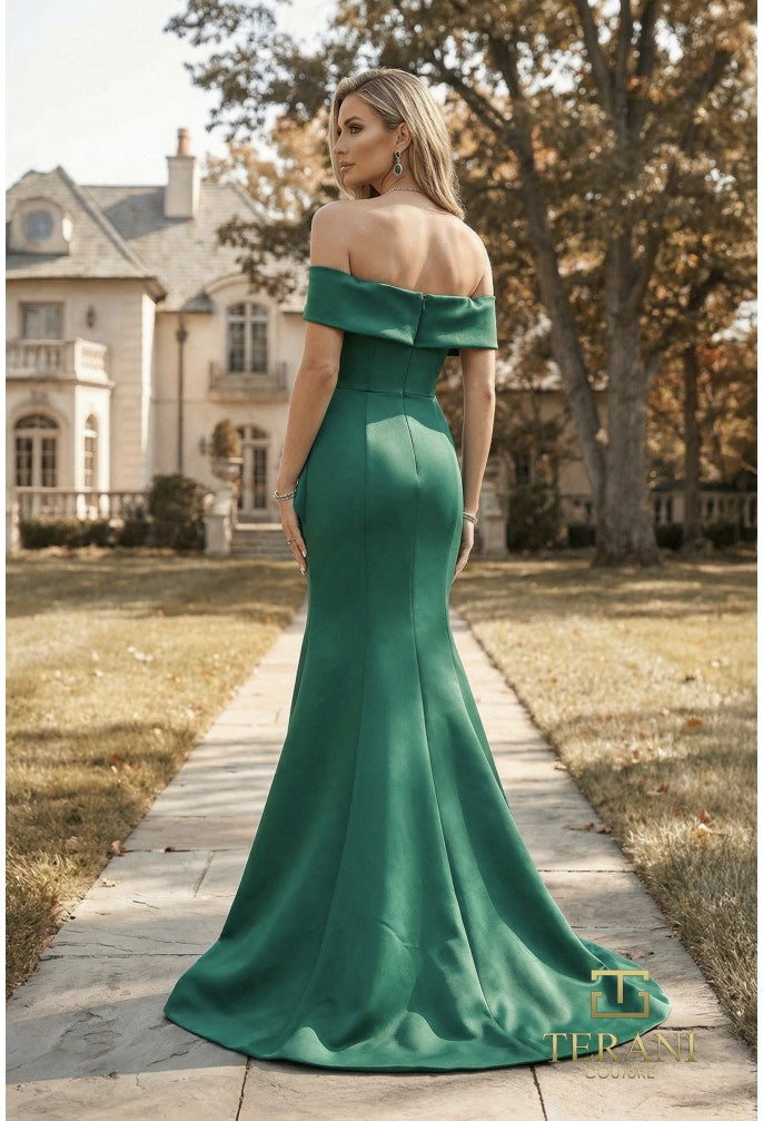 A woman stands on a stone path outdoors wearing an off-the-shoulder emerald green mermaid gown with a slight train. A large house and trees are visible in the background.