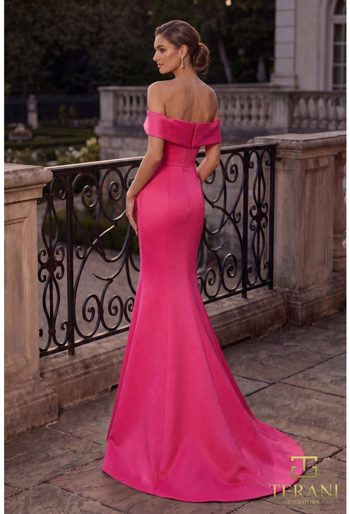 A woman models a fuchsia colored off-the-shoulder evening gown with floral embroidery on the bodice, standing on an outdoor stone terrace with a black wrought-iron railing. The Terani logo is visible in the bottom right corner.
