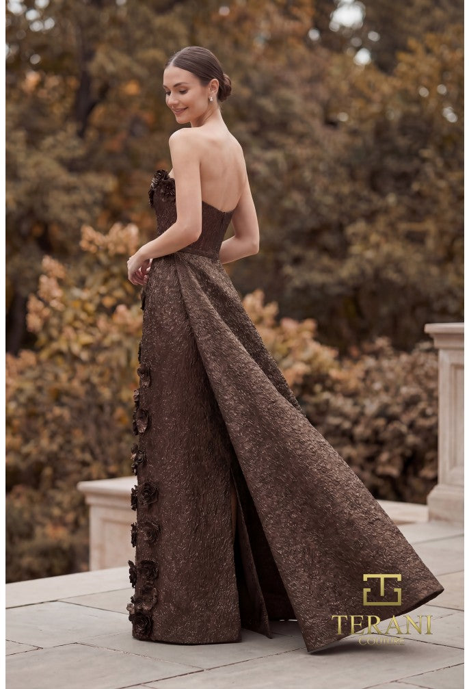 A woman wearing a strapless floor-length evening gown in a bronze color textured fabric, photographed outdoors on a stone terrace with trees in the background. The brand name TERRANT is visible in the lower right corner.