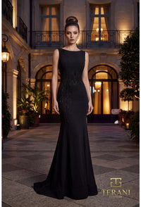 A woman stands in a grand courtyard wearing a floor-length black evening gown with a fitted silhouette, beaded embellishment along the side, and a slight train. The background shows illuminated windows and architectural details at night.