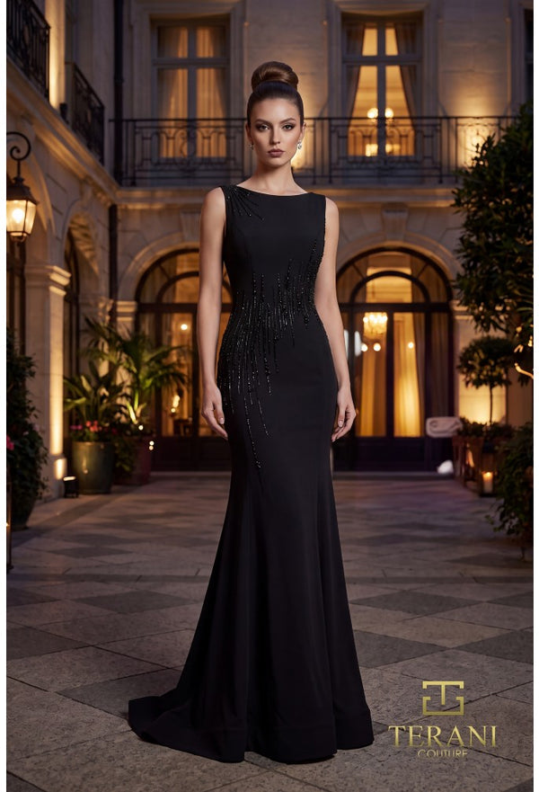 A woman stands in a grand courtyard wearing a floor-length black evening gown with a fitted silhouette, beaded embellishment along the side, and a slight train. The background shows illuminated windows and architectural details at night.