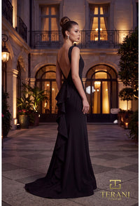 A woman modeling a floor-length black evening gown with a deep back and layered ruffle detail, standing in a courtyard at night with warm-lit building architecture in the background. The Terani Collection logo appears in the lower right corner.