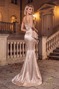 A woman models a champagne satin strapless mermaid gown with a structured bodice and dramatic draped side detail, posed outdoors in front of a grand stone building at night.