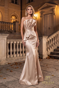 A woman models a champagne satin strapless evening gown with a draped bodice and flared skirt, standing on a stone terrace in front of a grand building at night.