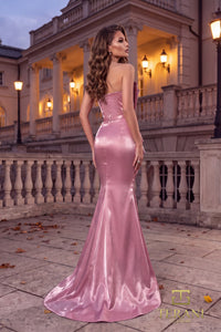 A woman stands on a stone terrace in front of an elegant building at dusk, wearing a floor-length satin mermaid gown in a rose pink color. The dress has a strapless backless design and a slight train.