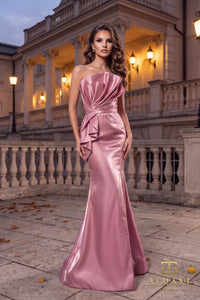 A woman models a floor-length, strapless satin gown in a rose pink shade, standing outdoors in front of a grand classical building at dusk. The dress features a structured, draped bodice with a ruffled overskirt and a fitted mermaid silhouette.