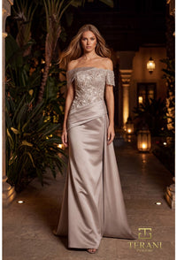 A woman models an elegant off-the-shoulder evening gown in a warm, tropical-themed hallway. The gown features a heavily embroidered lace bodice and a flowing satin skirt in a champagne-beige color.