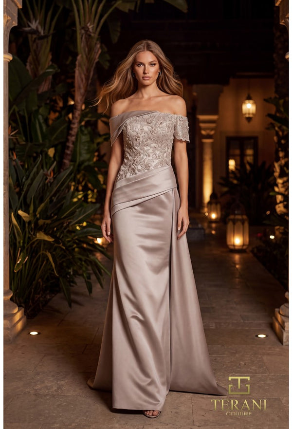 A woman models an elegant off-the-shoulder evening gown in a warm, tropical-themed hallway. The gown features a heavily embroidered lace bodice and a flowing satin skirt in a champagne-beige color.