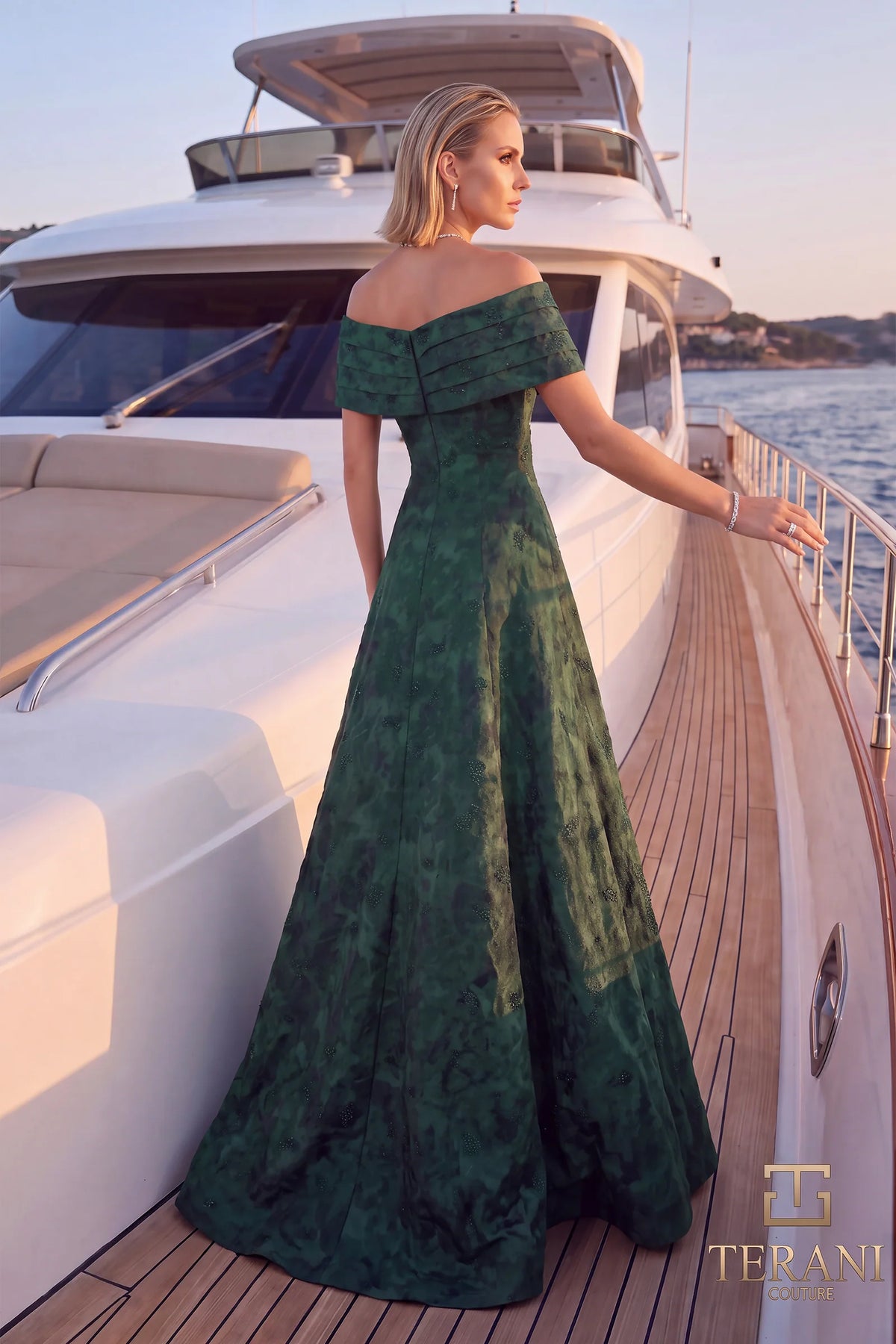 A woman stands on the deck of a luxury yacht wearing a floor-length off-the-shoulder evergreen green gown with a textured, marbled pattern. The Terani logo is visible in the bottom right corner.