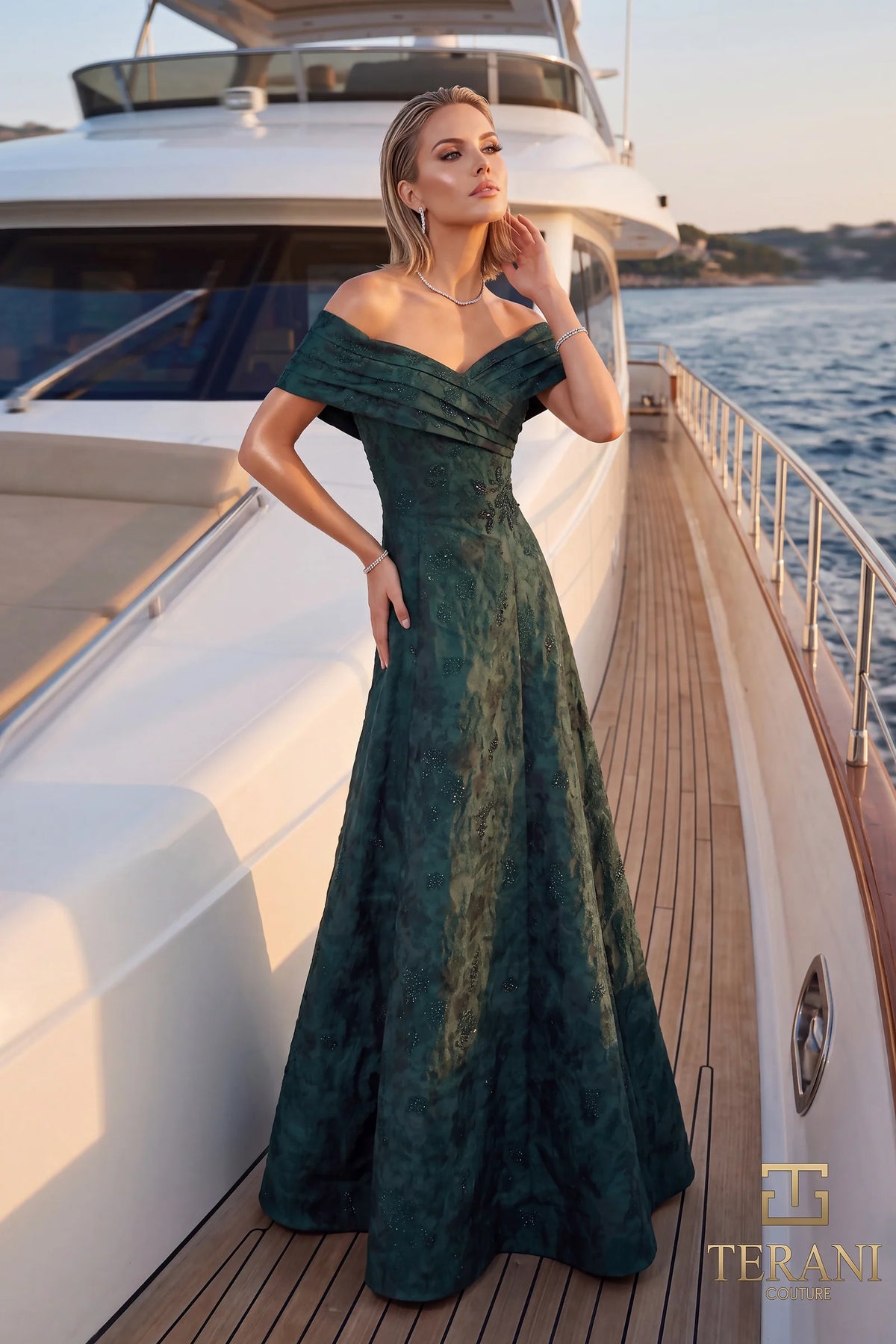 A woman models an off-the-shoulder floor-length gown on a yacht deck with water and sky in the background. The dress is a evergreen with a subtle textured, marbled pattern.
