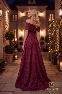 A woman wearing a floor-length off-the-shoulder merlot color gown with a textured jacquard pattern, standing in a stone courtyard at night with warm lights and potted plants.