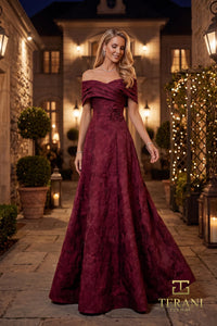 A woman wearing a floor-length off-the-shoulder merlot color gown with a textured jacquard pattern, standing in a stone courtyard at night with warm lights. The Terani Collection logo is visible in the bottom right corner.