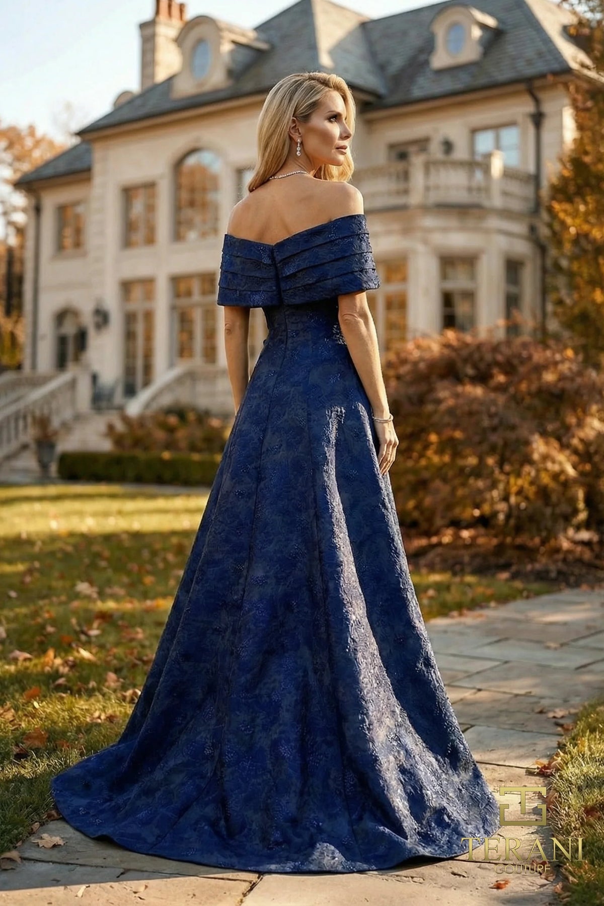 Woman in a midnight blue off-shoulder gown standing in front of a large house.
