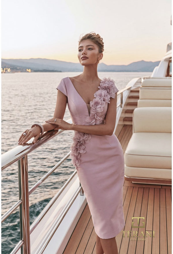 A woman models a light pink sleeveless dress with 3D floral appliqués on the bodice and skirt, posing on the wooden deck of a luxury yacht with water and mountains in the background.