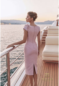 A woman stands on a yacht deck by the water at sunset, wearing a light pink knee-length dress with cap sleeves, a floral detail on one shoulder, and a back zipper. The brand name 'THE ANTI' is visible in the bottom right corner.