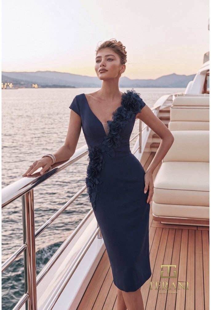 A woman models a navy blue knee-length dress with ruffle details on a yacht deck with water and mountains in the background.