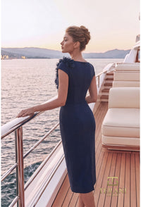 A woman wearing a navy blue knee-length dress with cap sleeves and a ruffled shoulder detail stands on a yacht deck, looking out over the water at sunset. The brand name 'THE RANA' is visible in the bottom right corner.