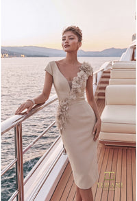 A woman models a beige knee-length dress with 3D floral appliqués on the bodice and one sleeve, posing on the deck of a luxury yacht with water and mountains in the background.