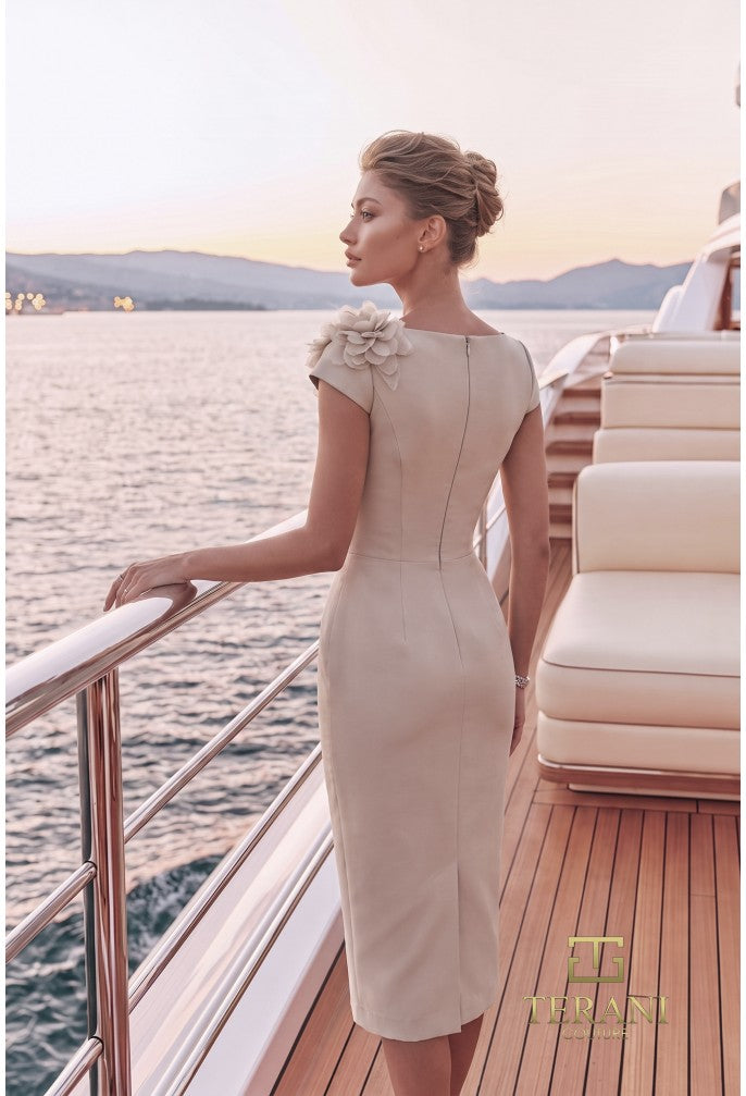 A woman stands on a yacht deck by the water at sunset, wearing a fitted beige knee-length dress with cap sleeves, a floral embellishment at the shoulder, and a back zipper. The brand name The Asani is visible in the lower right corner.